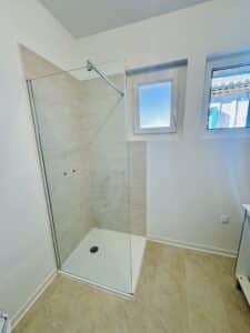 Douche à l'italienne moderne avec paroi en verre, carrelage beige et receveur blanc dans une salle de bain lumineuse.