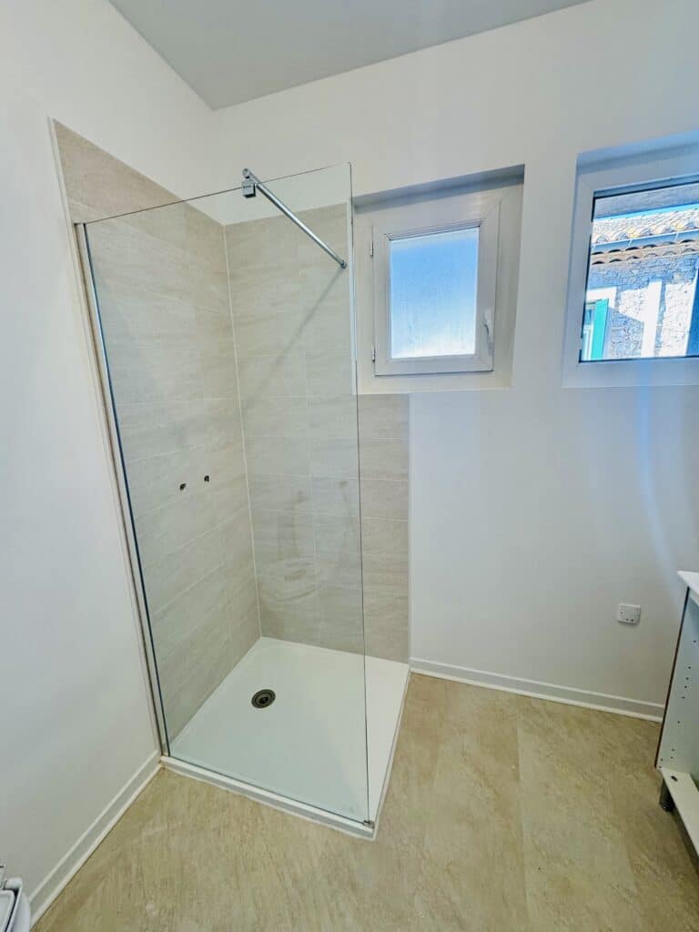 Douche à l'italienne moderne avec paroi en verre, carrelage beige et receveur blanc dans une salle de bain lumineuse.