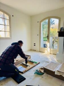 Artisan montant un meuble en bois avec une visseuse électrique dans un salon clair en cours d'aménagement.