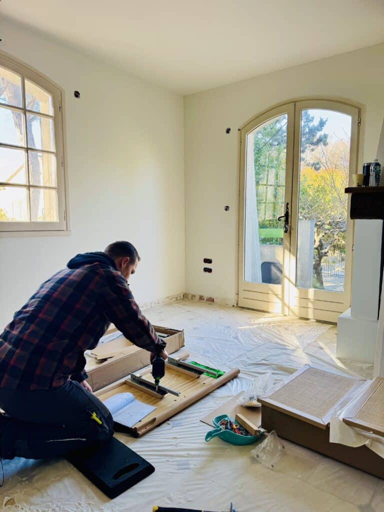 Artisan montant un meuble en bois avec une visseuse électrique dans un salon clair en cours d'aménagement.