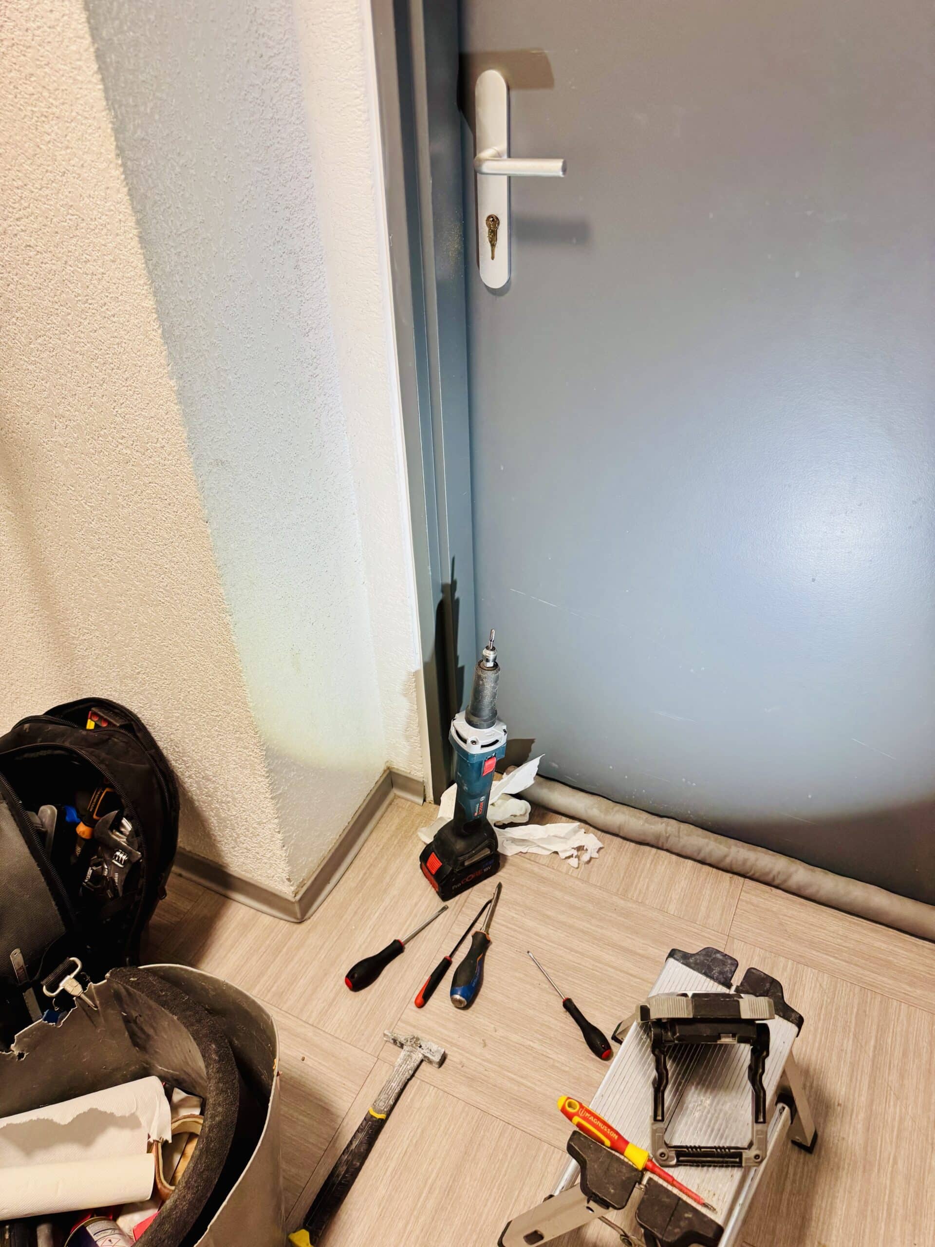 Outils de bricolage dont une perceuse Bosch ProCORE 18V, tournevis et marteau au sol devant une porte grise fermée.