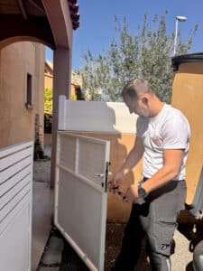 Technicien TVSB réparant le verrou d'un portillon blanc extérieur avec un tournevis.