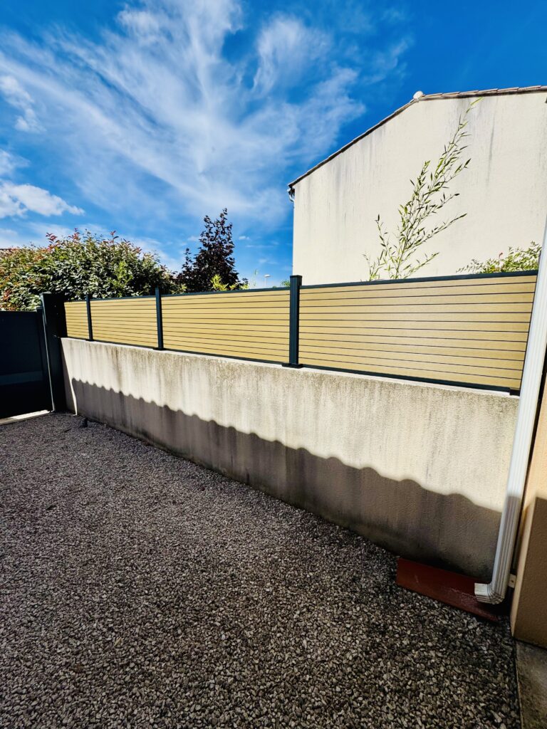 Clôture Composite sur Muret : Design Moderne et Durable Clôture composite bois et poteaux gris sur muret béton, jardin avec gravier sous ciel bleu.
