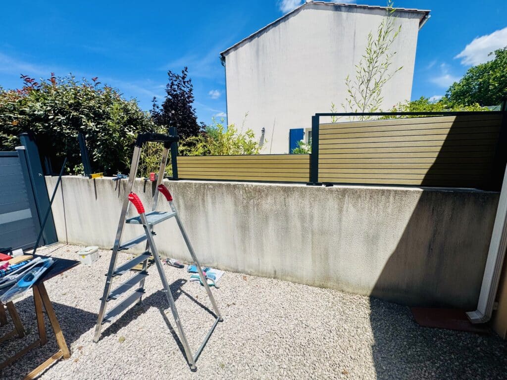 Échelle devant une clôture bois et gris sur mur en béton, jardin gravillonné sous ciel bleu.