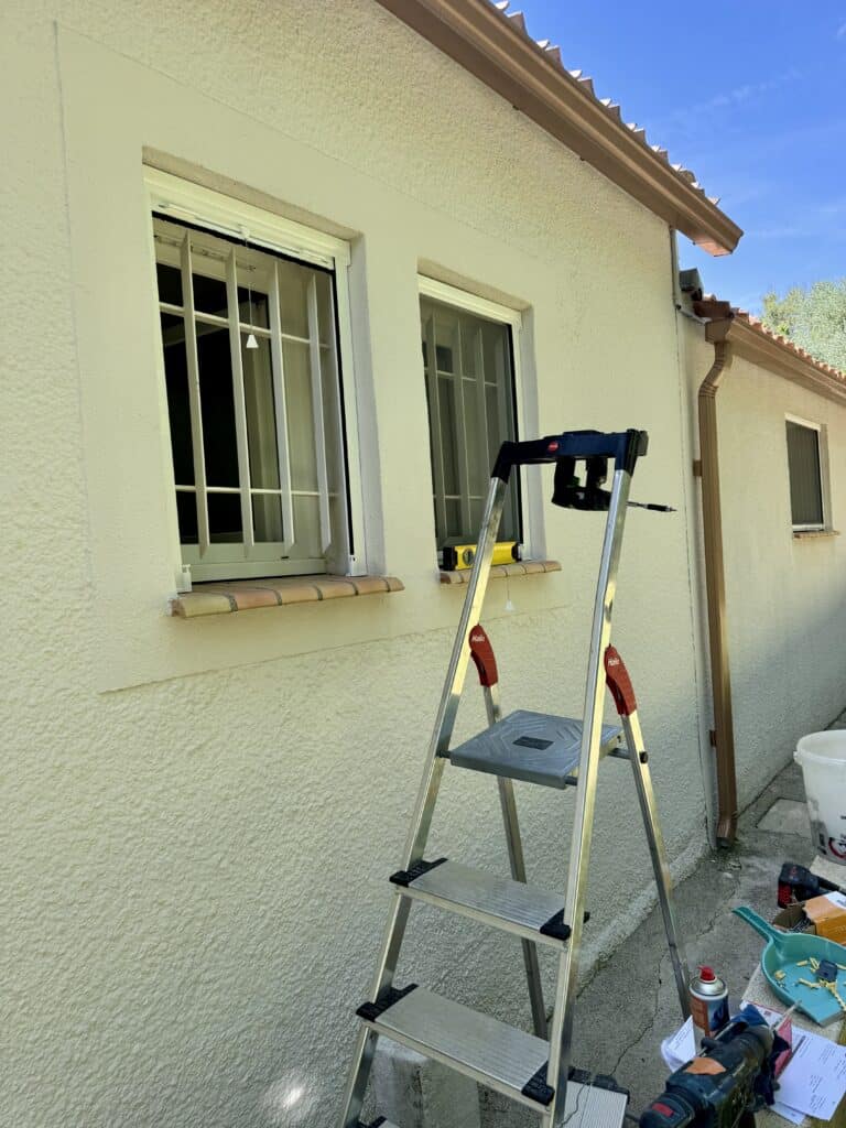 Installation grille sécurité fenêtre et échelle Hailo Échelle Hailo et outils devant une maison beige avec fenêtres sécurisées par des barreaux de protection.