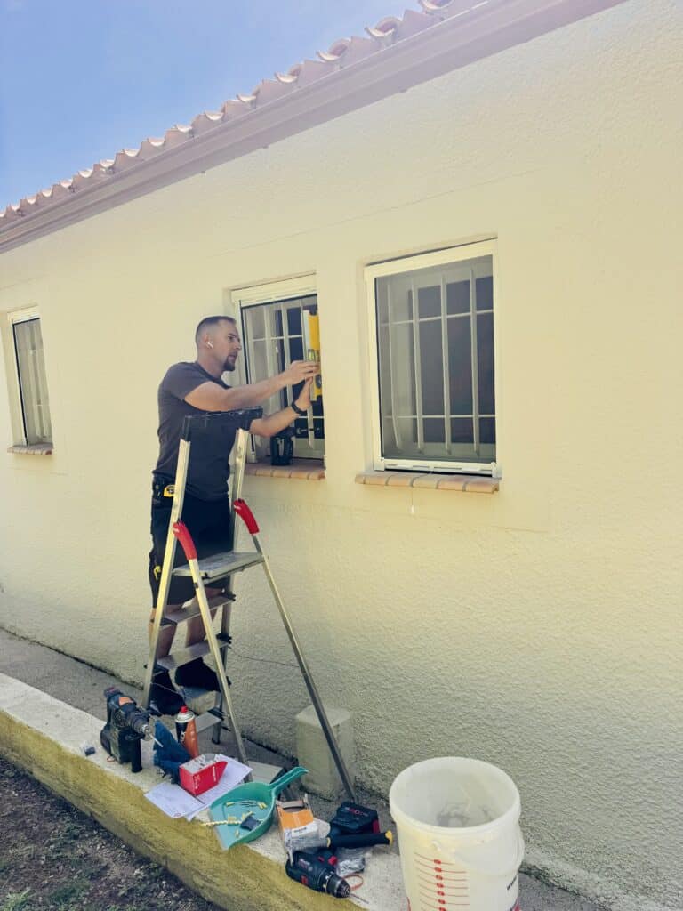 Pose de grilles de sécurité sur fenêtre : homme utilisant un niveau sur escabeau avec outils de bricolage.