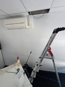 Maintenance climatiseur Mitsubishi avec escabeau Hailo sous un plafond technique ouvert.