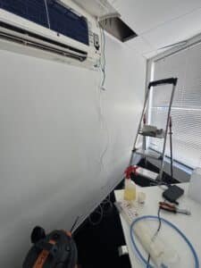 Maintenance d'un climatiseur Mitsubishi Electric dans un bureau avec échelle, outils et tubes de drainage.