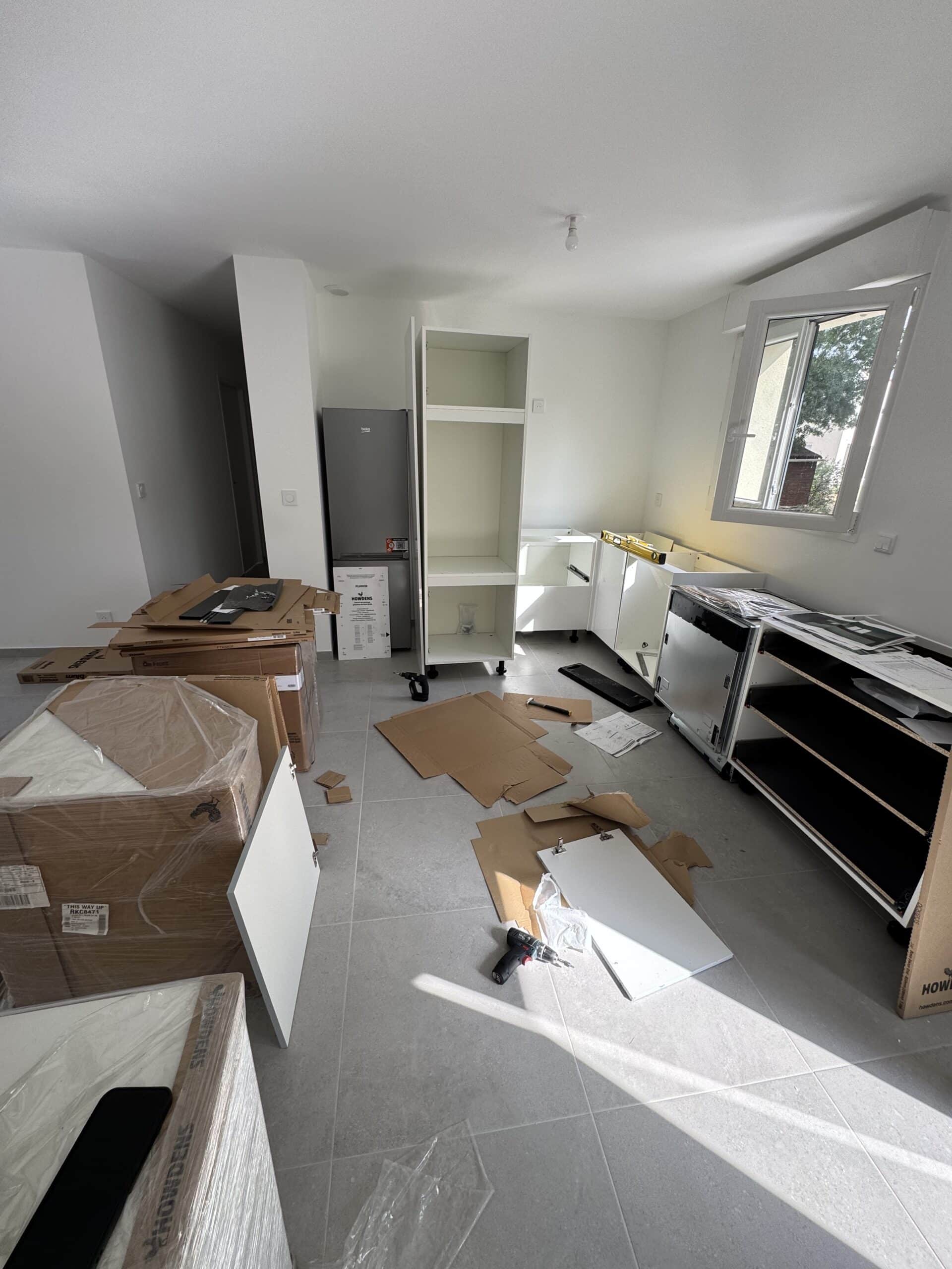 Cuisine Howdens en montage avec meubles blancs, outils et cartons sur carrelage gris.