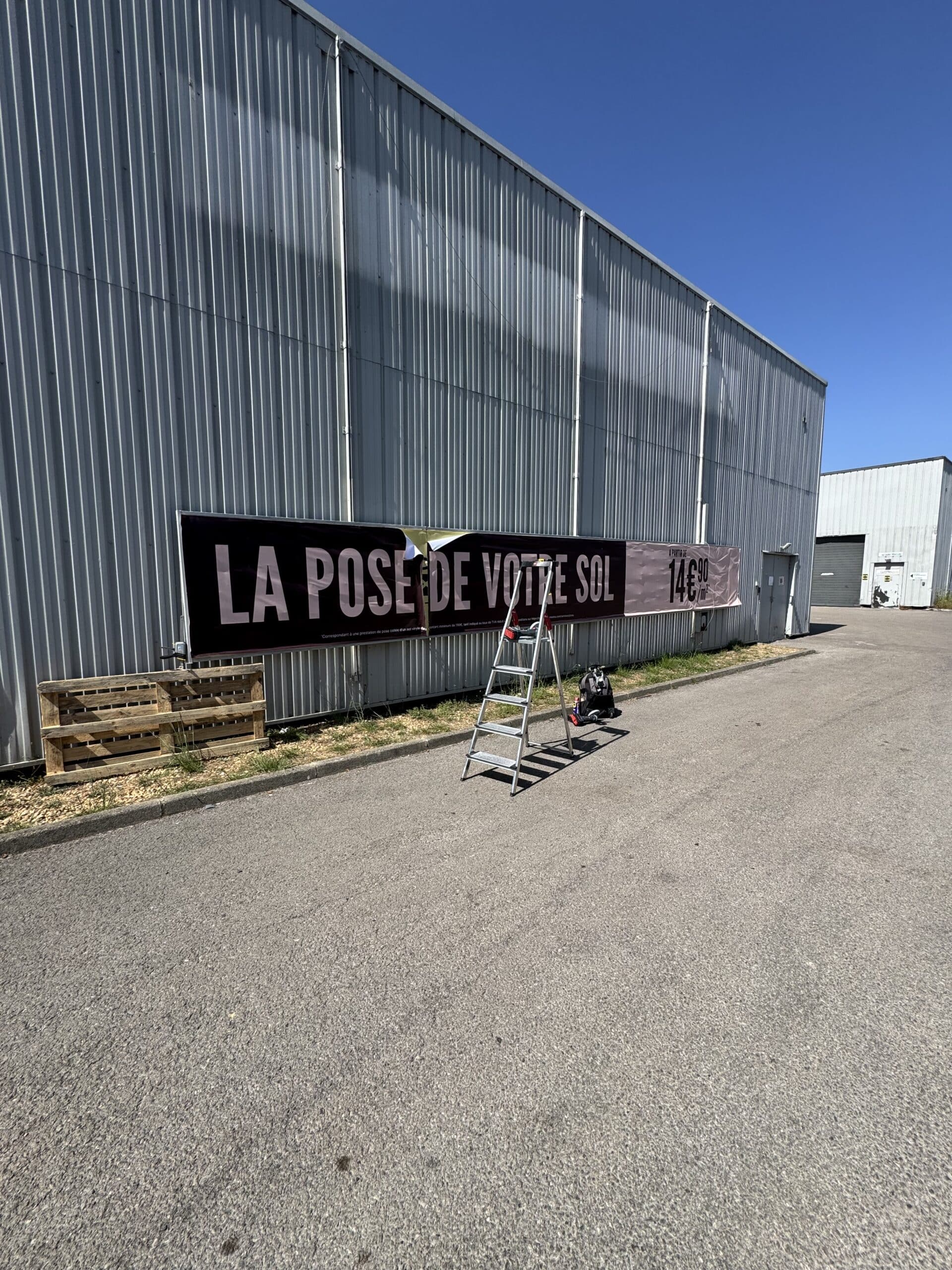 Bannière 'LA POSE DE VOTRE SOL 14€90' sur bâtiment industriel avec échelle et palette bois.