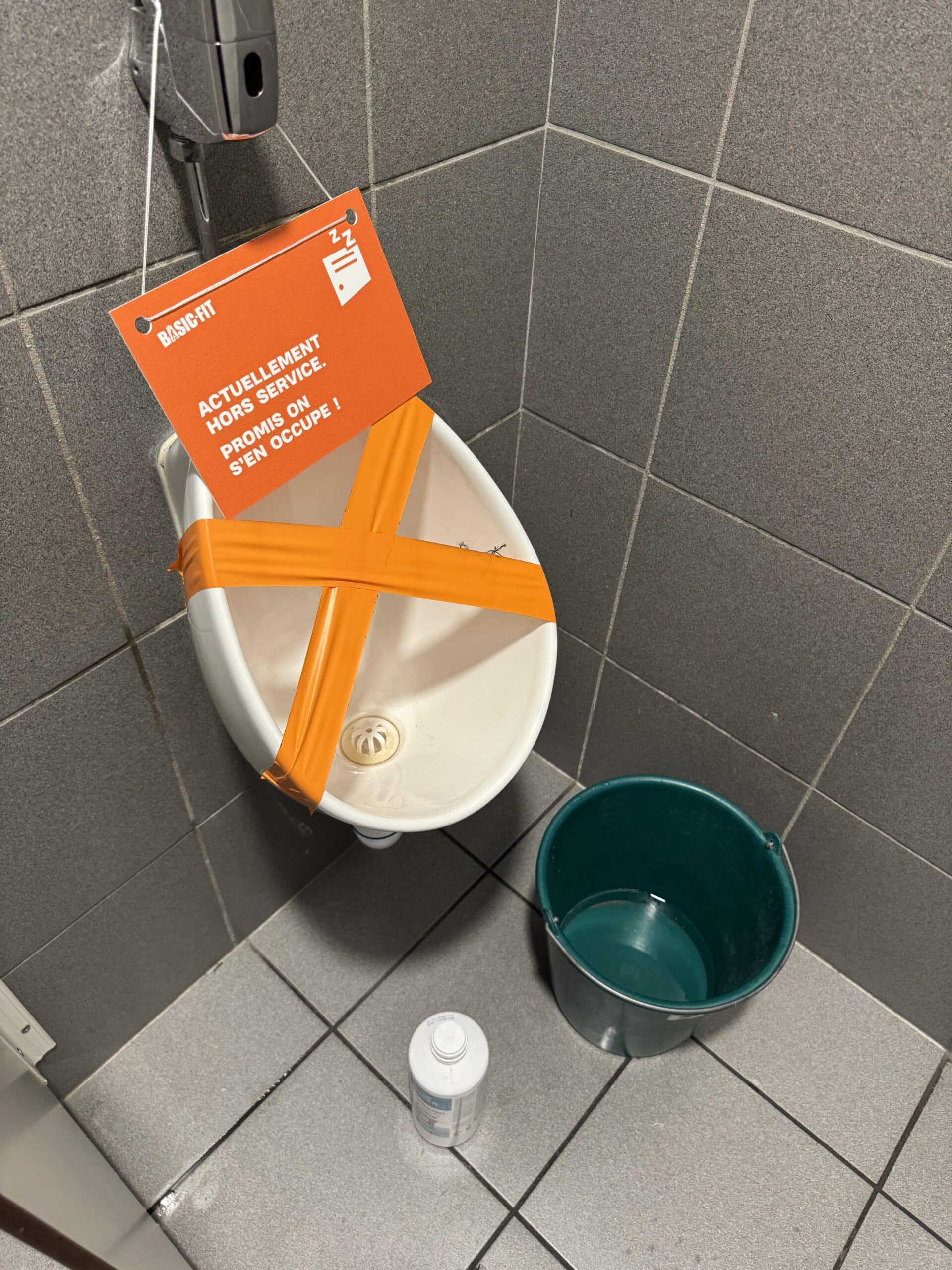 Urinoir Basic-Fit hors service avec ruban orange, panneau 'Promis on s'en occupe !', seau vert et flacon de nettoyage.